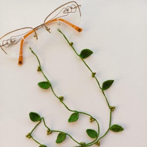 Op de afbeelding: Een bril met een oranje montuur en een groene gehaakte brillenketting. De ketting is versierd met groene bladeren en kleine kralen. De bril ligt op een wit oppervlak.