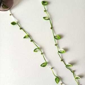 Op de afbeelding: Een zonnebril met een gehaakte brillenketting. De ketting is groen met kleine, gehaakte blaadjes en goudkleurige kralen. De ketting is bevestigd aan de zonnebril en ligt op een wit oppervlak.