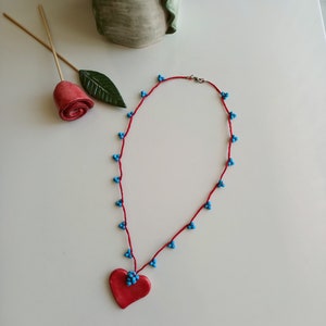 May include: A red heart-shaped pendant necklace with blue beads. The necklace is made of red string and has a silver clasp.