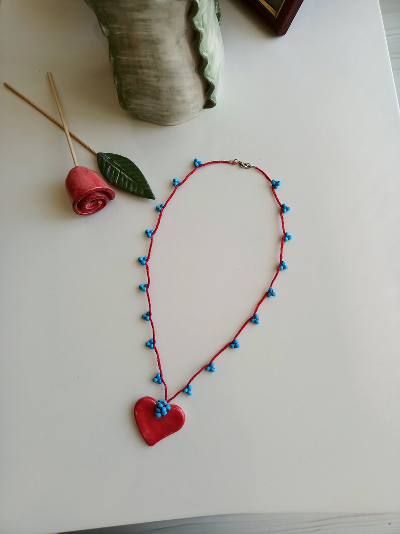 May include: A red heart-shaped pendant necklace with blue beads. The necklace is made of red string and has a silver clasp.
