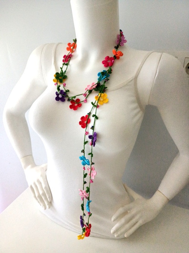 May include: A long, colorful crocheted necklace with small flower-shaped beads. The necklace is made of multiple strands of green yarn with colorful flowers in shades of red, orange, yellow, green, blue, purple, and pink.