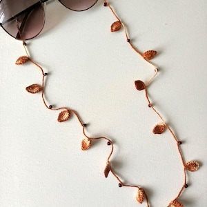 Op de afbeelding: Zonnebril met bruine verloopglazen rust op een handgemaakte gehaakte brillenketting. De ketting heeft kleine, bladvormige accenten in bruin- en beigetinten, afgewisseld met kleine, donkerbruine kralen. De ketting is ontworpen om brillen vast te houden.