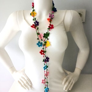 May include: A long, colorful crocheted necklace with small flower-shaped beads. The necklace is made with green, pink, red, blue, yellow, and purple yarn.