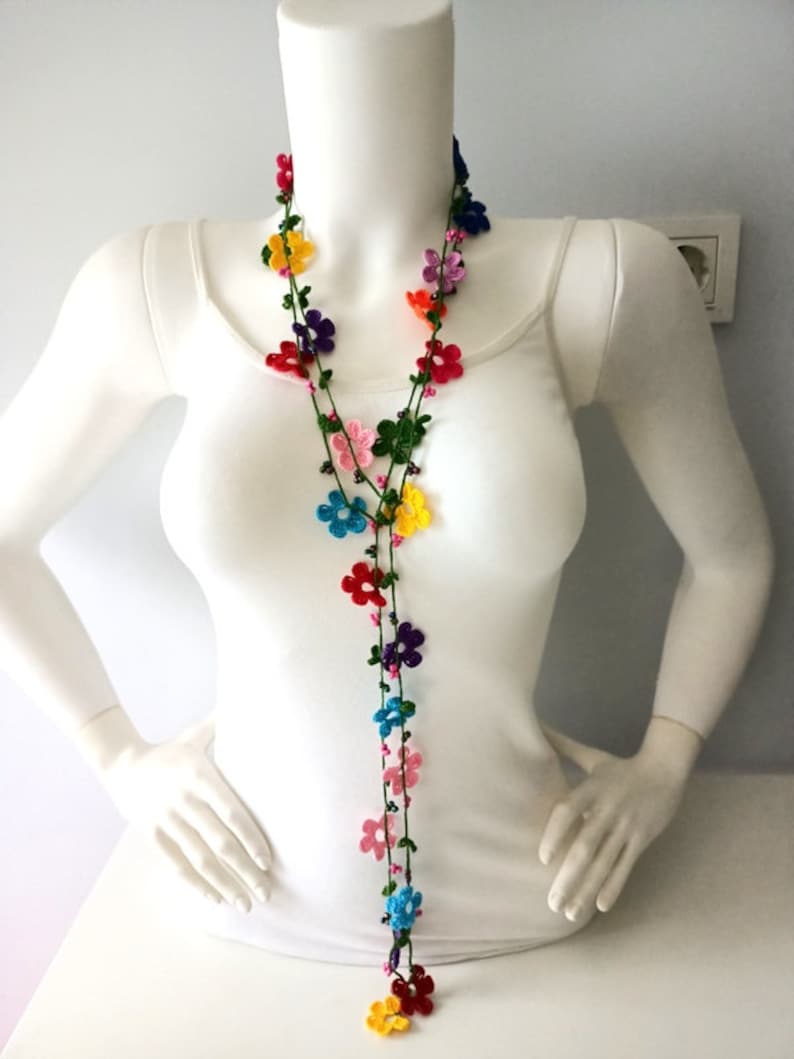 May include: A long, colorful crocheted necklace with small flower-shaped beads. The necklace is made with green, pink, red, blue, yellow, and purple yarn.