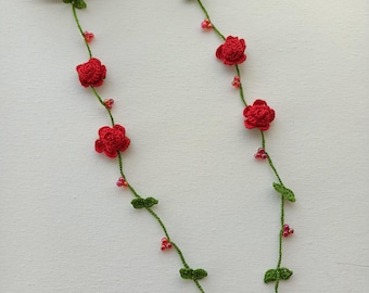 May include: A pair of black sunglasses attached to a green crocheted eyeglass chain featuring small red rose motifs, green leaves, and tiny red beads interspersed along the length of the cord.