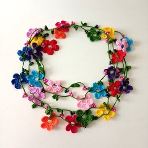May include: A multi-colored crocheted flower necklace with green leaves and black beads. The necklace is made of multiple strands of crocheted flowers in various colors, including pink, blue, yellow, orange, red, and purple.