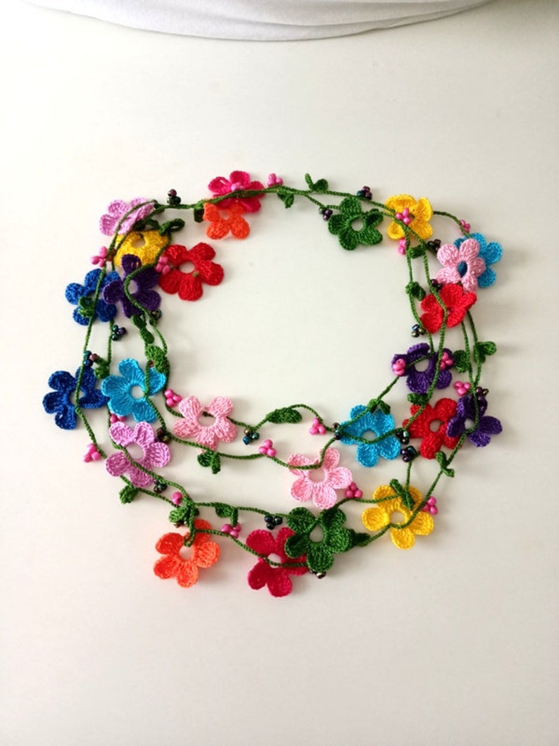 May include: A multi-colored crocheted flower necklace with green leaves and black beads. The necklace is made of multiple strands of crocheted flowers in various colors, including pink, blue, yellow, orange, red, and purple.