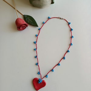 May include: A red necklace with a heart pendant. The necklace is made of red string with small blue beads. The heart pendant is also red.