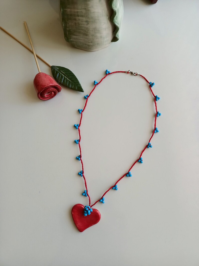 May include: A red necklace with a heart pendant. The necklace is made of red string with small blue beads. The heart pendant is also red.