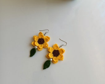 May include: A pair of handmade crochet sunflower earrings. Each earring features a yellow sunflower with a brown center, a green leaf, and a small white bead. The earrings have silver-colored fish hook ear wires.