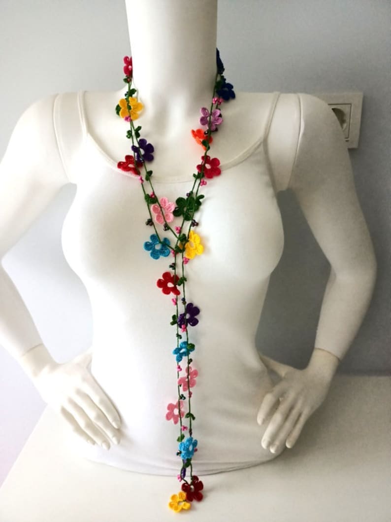 May include: A long, colorful crocheted necklace with small flower-shaped beads. The necklace is made with a variety of colors, including red, orange, yellow, green, blue, purple, and pink.