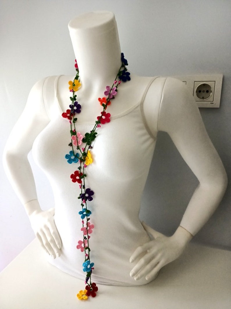 May include: A long, colorful crochet necklace with small flower-shaped beads. The necklace is made with green, pink, blue, yellow, red, and purple yarn.