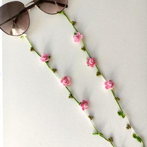 Op de afbeelding: Een bril met een gehaakte bloemenbrilkoord. De ketting is versierd met roze bloemen en groene bladeren.
