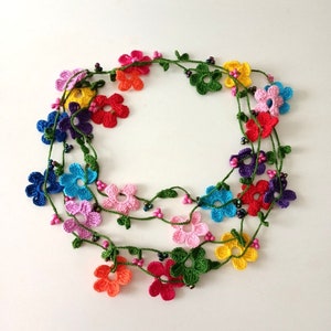 May include: A multi-colored crocheted flower necklace with green leaves and black beads. The necklace is made of multiple strands of crocheted flowers in various colors, including pink, red, orange, yellow, green, blue, and purple.