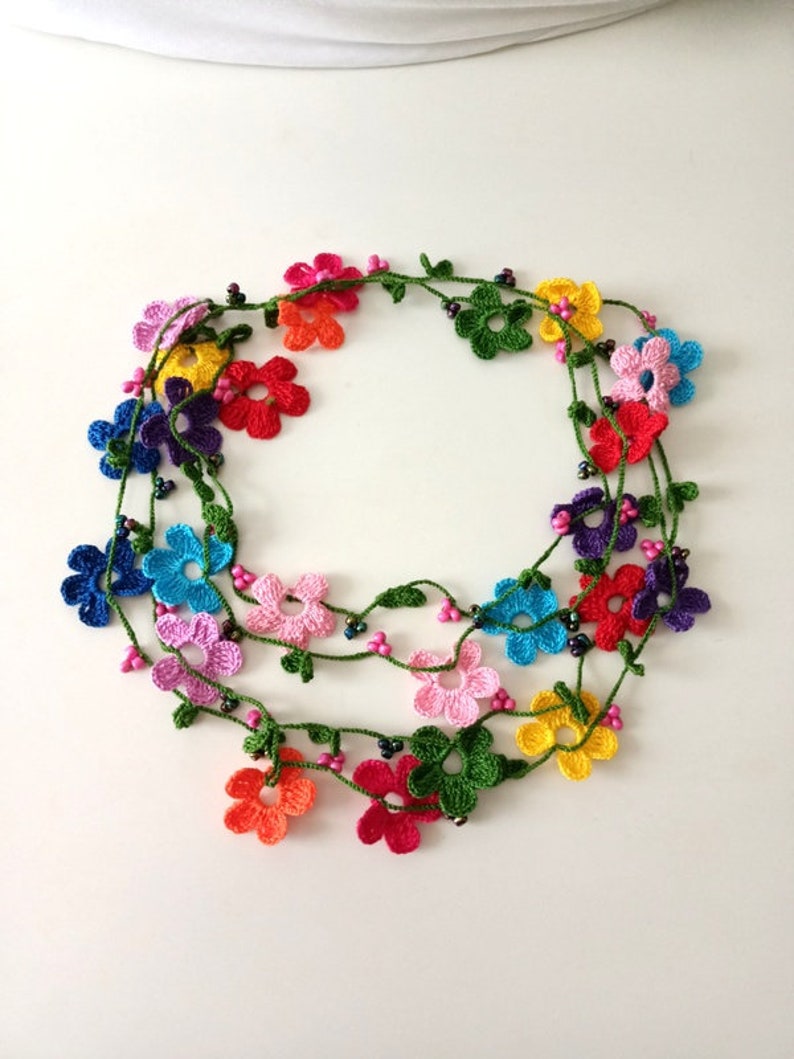 May include: A multi-colored crocheted flower necklace with green leaves and black beads. The necklace is made of multiple strands of crocheted flowers in various colors, including pink, red, orange, yellow, green, blue, and purple.