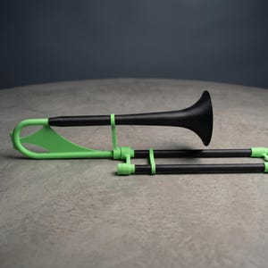 Könnte beinhalten: Eine Posaune mit einem schwarzen Korpus in Carbonfaser-Optik, einem ausgestellten Schallbecher und leuchtend hellgrünen Akzenten an Zug, Verstrebungen und Mundstückaufnahme. Das Instrument hat eine schlanke, moderne Bauweise.