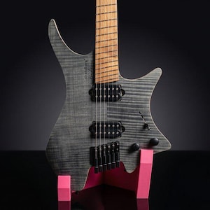 Può includere: Una chitarra elettrica headless con top in acero fiammato grigio, caratterizzata dalla scritta strandberg Boden sul corpo, due pickup humbucker neri, una tastiera in legno marrone chiaro e hardware nero, poggiata su un supporto in plastica rosa acceso.