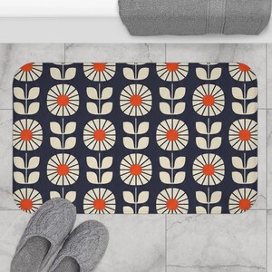 May include: A rectangular bath mat featuring a repeating pattern of stylized cream-colored flowers with bright orange centers on a dark navy blue background. The mat has rounded corners and a consistent floral motif arranged in vertical columns.