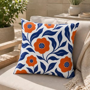 May include: A square throw pillow featuring a white background with a repeating pattern of vibrant orange flowers with dark blue centers and dark blue leafy stems. The design covers the entire front surface of the pillow in a consistent layout.