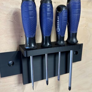 May include: A black wall-mounted tool organizer holding four screwdrivers with blue and black textured handles. One screwdriver is clearly branded with the word KOBALT. The organizer is secured to a light-colored wooden surface with visible screws.