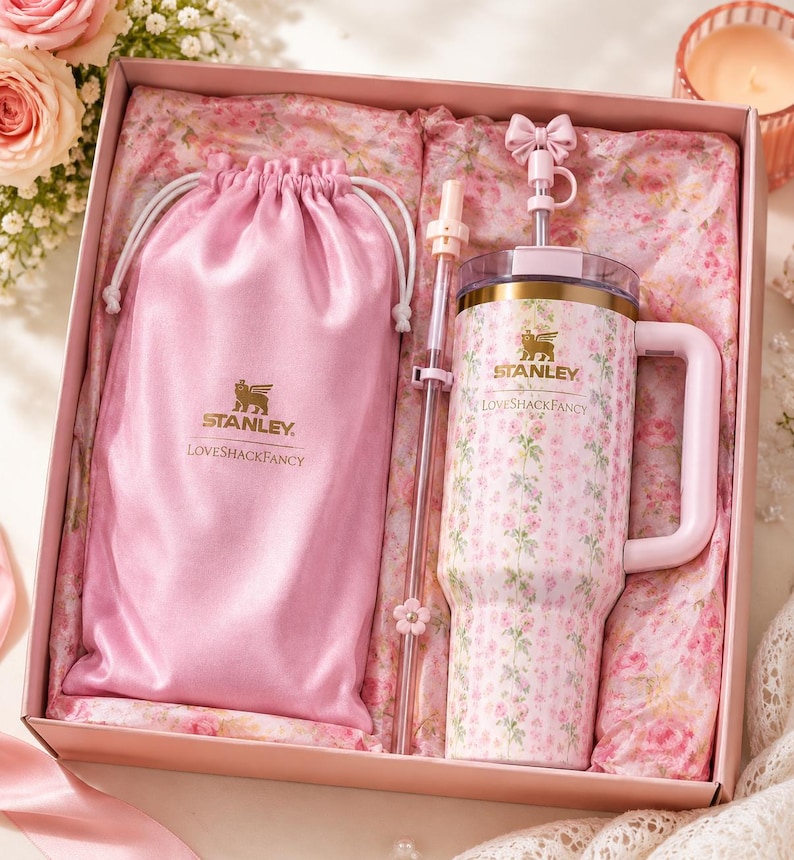 May include: A pink gift box containing a Stanley LoveShackFancy tumbler with a floral pattern, a matching pink satin drawstring pouch with the Stanley LoveShackFancy logo, and a clear straw featuring a small pink bow and flower charm topper.