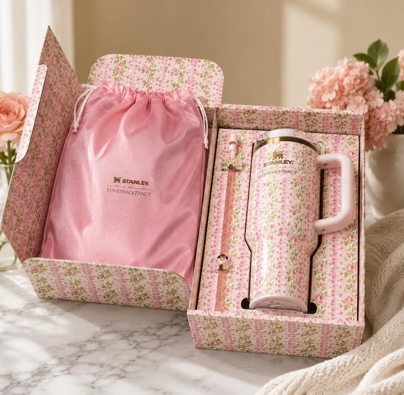 May include: A Stanley and LoveShackFancy collaboration set featuring a pink floral tumbler with a handle, a matching straw, and a pink satin drawstring bag. The items are presented in a floral patterned box with the Stanley and LoveShackFancy logos.