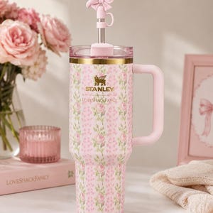 May include: A pink Stanley tumbler with a floral pattern, featuring the text Stanley and LoveShackFancy. The cup has a gold rim, a side handle, and a clear lid with a metal straw topped by a pink bow-shaped silicone straw cover accessory.