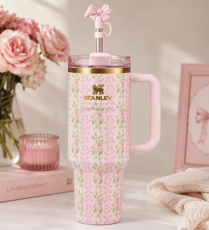 May include: A pink Stanley tumbler with a floral pattern, featuring the text Stanley and LoveShackFancy. The cup has a gold rim, a side handle, and a clear lid with a metal straw topped by a pink bow-shaped silicone straw cover accessory.