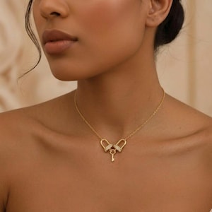 May include: A gold-toned necklace featuring a delicate chain and a central pendant composed of two interlocking U-shaped links adorned with small clear stones, with a small key charm suspended from the centre of the two links.