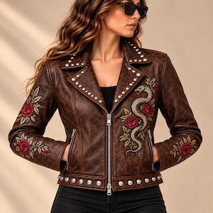 May include: A brown leather jacket featuring a repeating pattern, silver metal studs along the lapels and hem, and embroidered red rose and snake motifs on the front and sleeves. The jacket has a central silver zip and two zipped pockets.