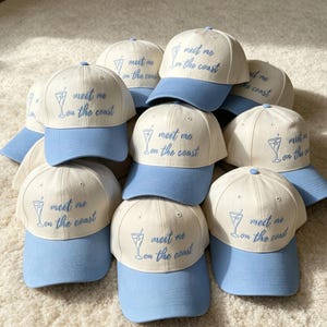 May include: A collection of baseball caps featuring a cream-colored crown and a light blue brim. Each cap is embroidered with a blue martini glass icon and the text "meet me on the coast" in a cursive script font across the front panel.