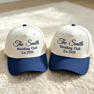 May include: Two off-white baseball caps with dark blue brims and top buttons. The front panels feature dark blue embroidered text that reads The Smith Wedding Club Est. 2026 in a combination of script and sans-serif fonts with ventilation eyelets.