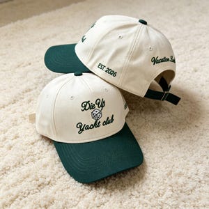 Puede incluir: Dos gorras de béisbol con coronas color crema y viseras verde oscuro. La gorra frontal presenta el texto bordado en verde oscuro Die Up Yacht club con un icono de dado. La gorra trasera muestra EST. 2026 y Vacation Land en verde oscuro.