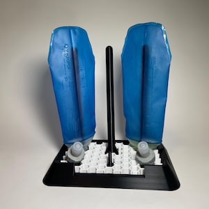 Op de afbeelding: Een zwart-wit kunststof droogrek met een centrale verticale paal en een basis met honingraatpatroon. Twee blauwe HydraPak-hydratatiereservoirs zijn ondersteboven op het rek geplaatst, met de grijze schroefdoppen naar voren gericht.