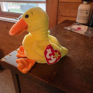 May include: A yellow plush duck toy with an orange beak and orange webbed feet. The stuffed animal features a small black eye and a heart-shaped red Ty Beanie Baby tag attached to its side with a plastic fastener, displaying the white Ty logo.