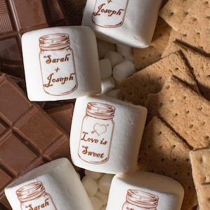 May include: A close-up of five white marshmallows with brown printed designs. The marshmallows are arranged on a burlap surface with chocolate squares and graham crackers. The designs on the marshmallows include a mason jar with the text "Sarah + Joseph" and a mason jar with the text "Love is Sweet".