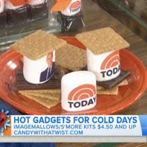 May include: A plate of s'mores kits with graham crackers, chocolate, and marshmallows. The marshmallows are branded with the "TODAY" logo and topped with graham crackers. The plate is orange and the background is blurred.