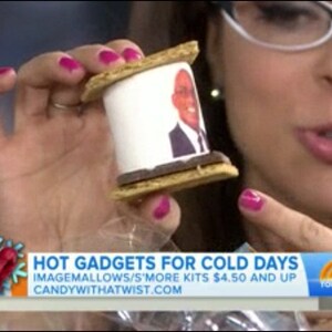 May include: A s'more featuring a white marshmallow with a printed image, sandwiched between graham crackers and a chocolate layer. The image is held by a person with pink nail polish. The image is part of a segment called "Hot Gadgets for Cold Days".