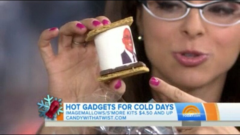May include: A s'more featuring a white marshmallow with a printed image, sandwiched between graham crackers and a chocolate layer. The image is held by a person with pink nail polish. The image is part of a segment called "Hot Gadgets for Cold Days".