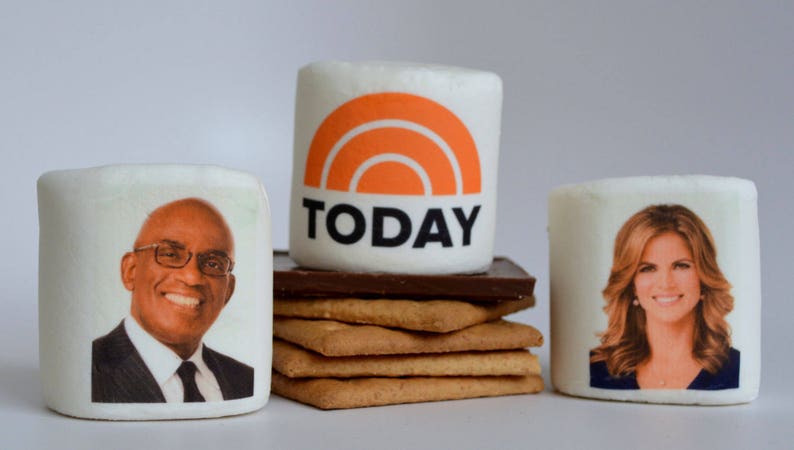 May include: Marshmallows with images of Al Roker and Natalie Morales, and the Today show logo. The marshmallows are stacked on graham crackers and chocolate, resembling a s'more.