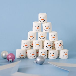 May include: A pyramid of white marshmallows with orange carrot noses and black eyes, arranged to look like snowmen. There are two small ornaments, one silver and one pink, in the foreground.