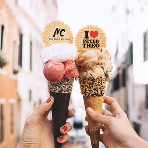 May include: Two ice cream cones are held up in front of a blurred background. One cone has pink and white ice cream with a waffle cookie that says "NC Visit North Carolina". The other cone has brown ice cream with a waffle cookie that says "I ❤️ PETER THEO".