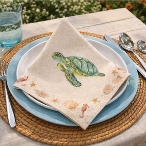 May include: A square fabric napkin featuring a watercolor illustration of a green sea turtle centered on a sandy beige background. The border is decorated with small orange starfish, seashells, and seahorses arranged around the edges.