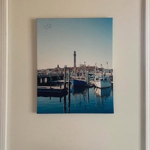 May include: A painting depicting a harbour scene with boats moored at a pier. The boats are blue and white, with a tall tower visible in the background.