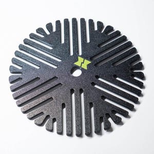 May include: A circular black disc with a textured finish featuring a radial pattern of cut-out slots and a central hole. A small, lime green logo shaped like two mirrored triangles is printed near the center of the disc, which has a symmetrical design.