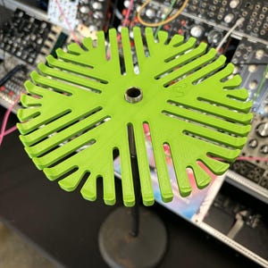 May include: A lime green circular cable management rack with numerous radial slots for organizing cords. The rack features a central metallic threaded insert and is mounted on a thin black vertical pole with a circular base for stability.