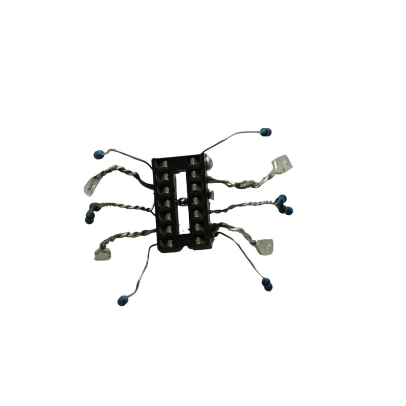 May include: A black rectangular electronic component socket with two parallel rows of pin slots. Several thin, twisted silver-coloured metal wires extend outward from the sides, each tipped with a small blue bead or a clear, translucent rectangular component.