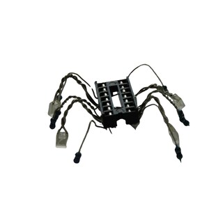 May include: A black electronic integrated circuit socket with eight twisted wire legs extending outwards. Each wire leg features a small, clear plastic component at the tip, resembling a mechanical spider-like structure made from electronic parts.
