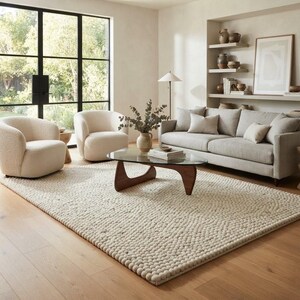 May include: A living room set featuring a light beige sofa with matching throw pillows, two cream-colored rounded armchairs, and a triangular glass-top coffee table with a dark wood base, all arranged on a large, textured, cream-colored area rug.