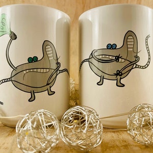 Op de afbeelding: Twee witte keramische mokken met een grillig cartoonontwerp. Elke mok heeft een grijs, gestileerd wezen met groene of blauwe ogen en een lange, gesegmenteerde staart. Eén houdt een gloeilamp vast. Decoratieve zilveren lichtslingers staan op de voorgrond.